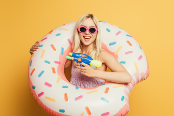 smiling blonde girl in swim ring holding water gun on yellow background