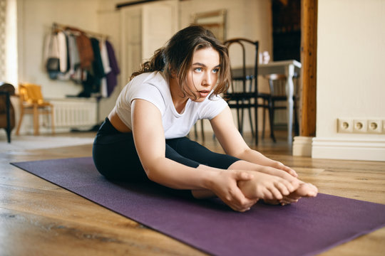 Concentrated young woman with flexible body sitting on mat with legs straight, doing seated forward bend or Paschimottanasana, leaning to her knees, keeping toes pointed. Cute girl stretching at home