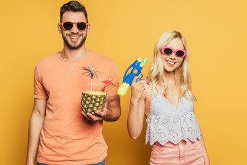 happy man with cocktail in pineapple and cheerful blonde girl with water gun on yellow background