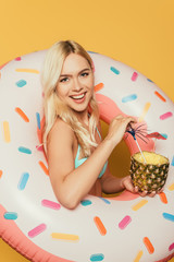 Obraz na płótnie Canvas happy blonde girl in swim ring holding half of pineapple with fresh cocktail on yellow background