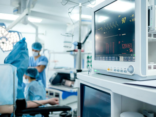 Monitoring the patient's vital functions in the operating room is in focus. Against the background of a strong blur, surgeons performing a surgical operation in the bright light of operating lamps.