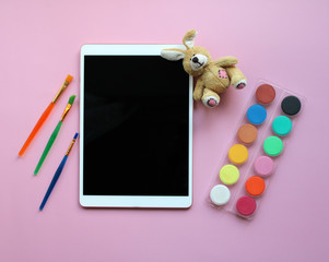 A tablet computer next to soft toy rabbit, multi-colored paints and art brushes are on a pink background. Children's creativity concept