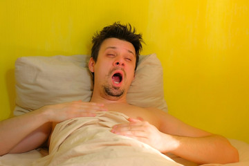 Sleepy bearded man with hangover wakes up, yawns in bed, portrait closeup. Young man with headache in the morning after waking up. Tired sick guy. Yellow wallpaper background.