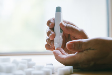 senior woman hands using lancet on finger at home to check blood sugar level, glucometer and sugar cubes on wooden table close up, diabetes concept, elderly health care, sunny morning