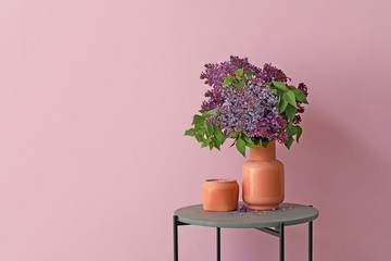 Vase with beautiful lilac flowers and candle on table against color background