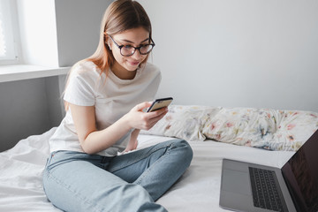 Obraz premium Young woman uses smartphone and laptop at home on the bed. Communication, video call, remote work