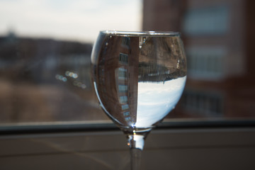 A glass of water on a thin leg stands on the windowsill. Interesting refraction of the view from the window. Sky and clouds in a glass. Optical illusion.