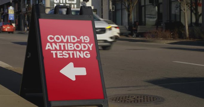 A sign on a sidewalk of a large city shows pedestrians and passing cars where the local COVID-19 antibody testing center is.	