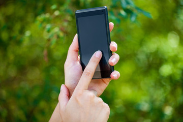 Side view of a woman's hand holding a modern slick smartphone while dialing with her thumb against summer green background