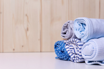 Obraz na płótnie Canvas Clothes stack for newborn boy. Close up of rolled up kid's chlothes on white chest of drawers in front of wooden wall. Grey and blue colors and different print. Copy space.