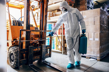 Man in protective suit and mask disinfecting forklift in warehouse from corona virus / covid-19.