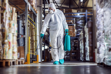 Man in protective suit and mask disinfecting warehouse full of food products from corona virus /...