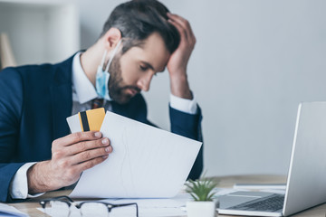 selective focus of depressed businessman holding paper and credit cards while sitting at workplace with bowed head and closed eyes