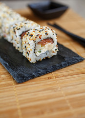 Uramaki Maki Sushi, Original roll served on a black board with black chopsticks and soy sauce in a black bowl.