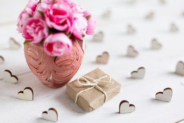 Beautiful composition with gift wrapped in kraft paper and delicate roses in pink vase on white wooden background