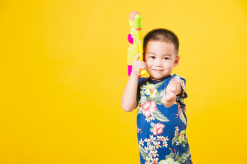 Portrait happy Asian cute little children boy smile standing so happy wearing flower shirt in Songkran festival day holding water gun, studio shot on yellow background with copy space