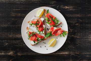 Sandwich with salmon, cheese, arugula and lemon on a white plate on a black table