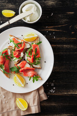 Sandwich with salmon, cheese, arugula and lemon on a white plate with a linen napkin and ingredients on a black table