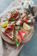 Sandwich with salmon, cheese, arugula and lemon on a wooden board with a linen napkin and ingredients on a gray table