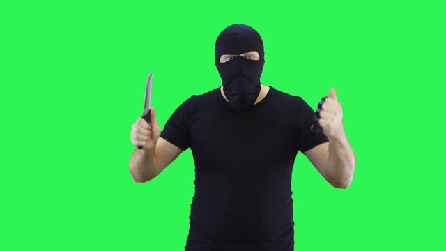 A man in a black mask holds a knife and brass knuckles in his hand, attacks.Balaclava.Green screen background.