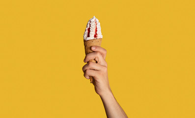 Millennial guy holding tasty ice cream with fruit topping in waffle cone, orange background, closeup. Panorama