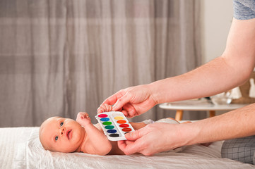 Naklejka premium Newborn with paints on a white plaid closeup. Infant holds watercolor rainbow paint and copy space. The concept of the game, care and care for the baby. 