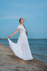 Fototapeta na wymiar Beautiful young woman in long white dress standing on sandy beach on sea background. Vertical frame