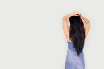 Beautiful young woman with long straight dark hair in blue silk nightdress on grey monotone background wall. Female turned back and put her hands on her head. Copy space for text and design.