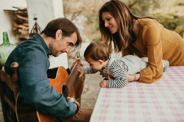 One year baby touching his father’s guitar while he plays at home courtyard. Mother is holding the baby. - obrazy, fototapety, plakaty