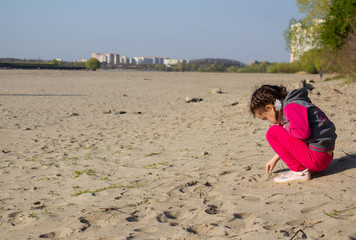 A little girl sits on the sand on the river Bank and touches the sand