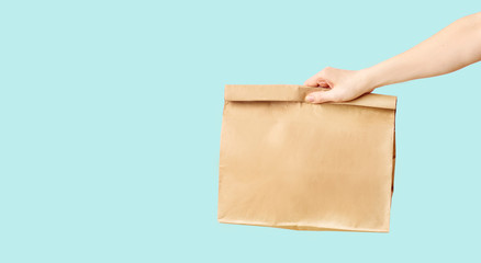 Female hand receiving brown clear empty blank paper bag for food delivery on blue background with copy space. Packaging template mock up fast food.