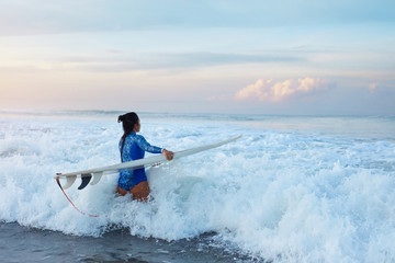 Fototapeta na wymiar Surfer Girl. Surfing Woman With Surfboard Going To Surf In Ocean. Brunette In Blue Wetsuit Walking Into Splashing Sea. Water Sport For Active Lifestyle.