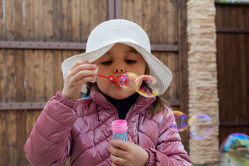A charming little girl in a white hat blows bubbles against the background of a wooden wall