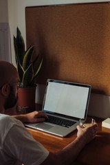 Man sitting at the table of his home office working on a laptop. Work from home concept. White blank space on the laptop's screen, space for text, copy space on the cork board