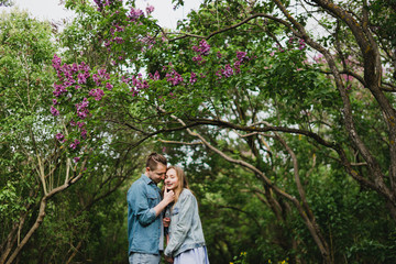 Romantic and happy caucasian couple in casual clothes hugging on the background of beautiful blooming lilac. Love, relationships, romance, happiness concept. Man and woman walking outdoors together.