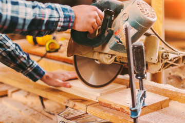 Closeup of skilled man carpenter using electric circular saw at woodworking workshop. Professional cabinet maker cutting wooden board with circuit saw at sawmill. Wooden furniture production factory