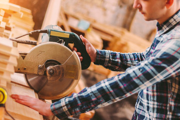 Closeup professional carpenter hold electric circular saw at sawmill. Skilled joiner using circular saw for cutting wood board at woodworking workshop. Furniture production factory. Man cabinet maker