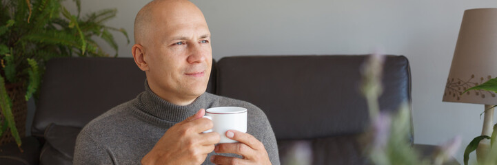 A satisfied man sits on a sofa at home and drinks coffee.