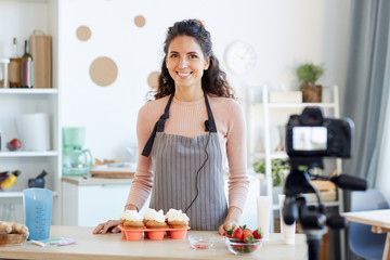 Portrait of young adult woman standing at kitchen table with homemade cupcakes with buttercream on it