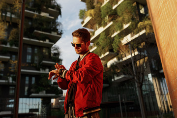 Hipster guy wear in cool clothes walking on the street looking at watch, in day time, buildings backgrounds.