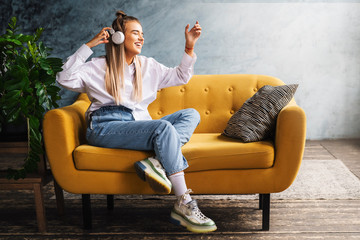 Obraz premium Young beautiful woman listens to music in headphones. Girl in white shirt sits on couch enjoys streaming audio, podcasts
