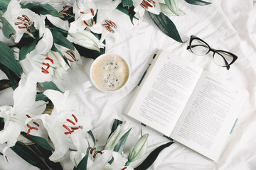 Opened book, coffee cup, glasses, fresh flowers on the white bed. Flat lay, top view