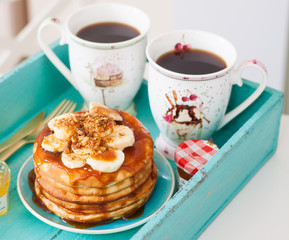 Stack of pancakes with maple syrup, chopped bananas and pecan nuts. Two cups of coffee on breakfast tray.