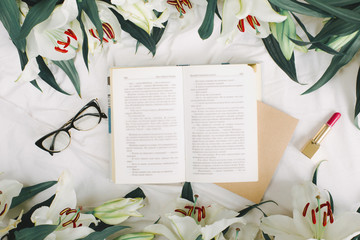 Opened book, red lipstick, glasses and fresh flowers on the white bed. Flat lay, top view