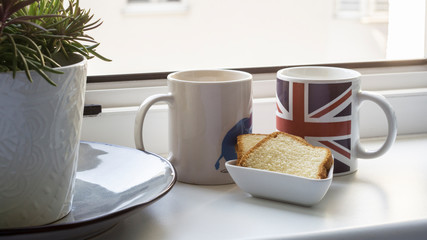 Two cups, two pieces of lemon cake and a plant in front of an open window. The picture shows love an togethernes