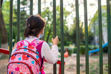 Obraz premium Little school girl sitting next to school fence waiting for going back to clases after pandemic outbreak