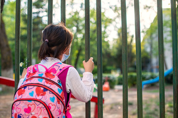 Little school girl sitting next to school fence waiting for going back to clases after pandemic outbreak