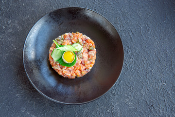 Fresh salmon tartar with quail egg, cucumber, greens on black table, textured dark table. Seafood raw concept. Copy space.