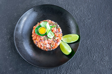 Fresh salmon tartar with quail egg, cucumber, greens on black table, textured dark table. Seafood raw concept. Copy space.
