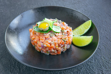 Fresh salmon tartar with quail egg, cucumber, greens on black table, textured dark table. Seafood raw concept. Copy space.